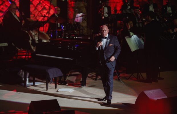 Michael Martocci performing on stage with a microphone in hand, accompanied by an orchestra including a pianist and brass instrumentalists, in a setting with red lighting and elegant decor.