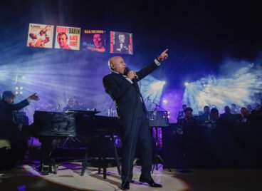 Michael Martocci performing on Altos de Chavón amphitheater with a microphone in hand, backed by an orchestra, under blue stage lights, with banners featuring Frank Sinatra, Louis Armstrong and Bobby Darin in the background.